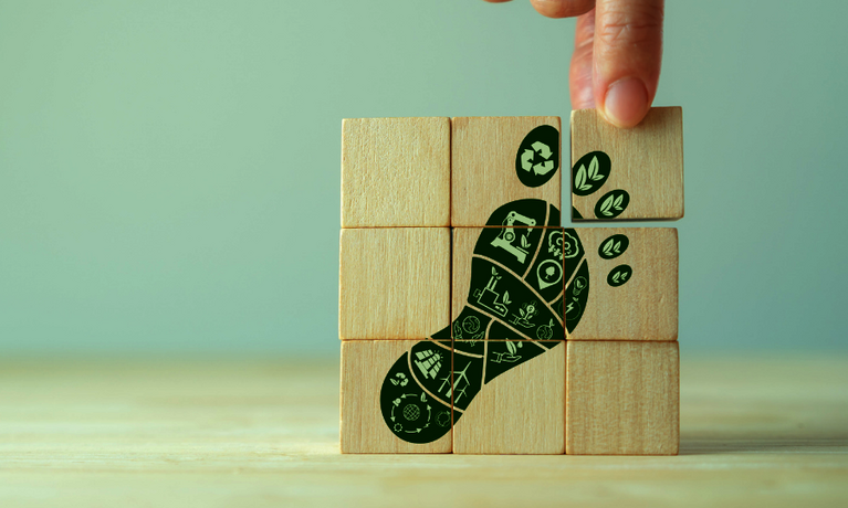 Neun Bauklötze mit dem Motiv eines grünen Fußabdrucks wie ein Puzzle zu einem Quadrat aufgebaut. Ein Daumen und Zeigefinger greift nach einem Würfel.