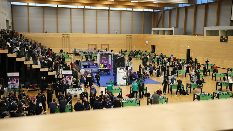 Halle mit mehreren Menschen die am Tischkicker ihr Glück versuchen. Auf der linken Seite tobende Menschen.